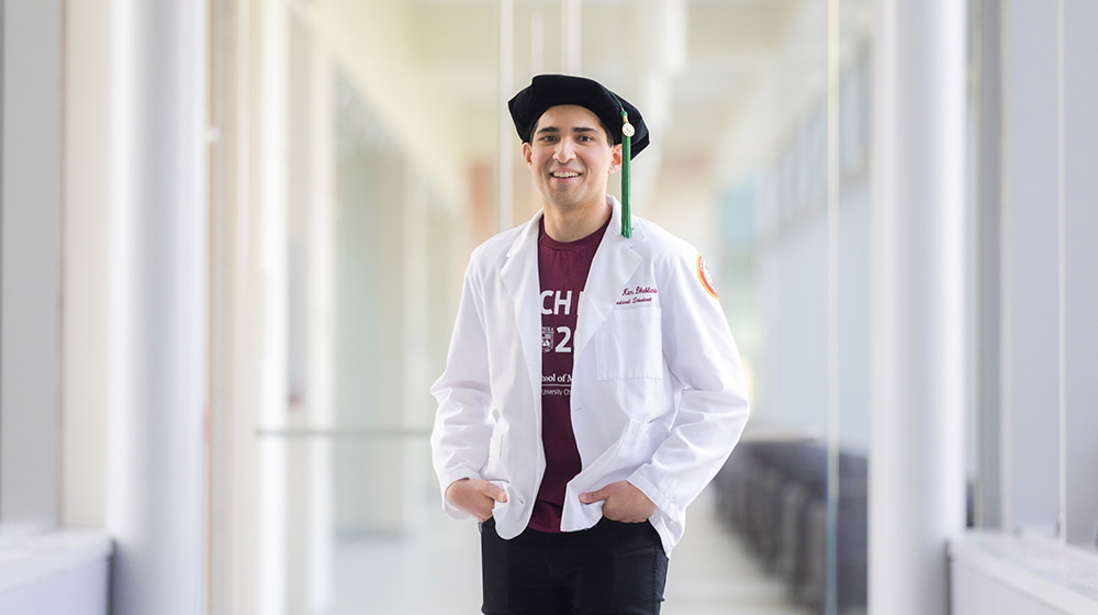 Nathan Dhablania standing in a hallway wearing maroon t-shirt, white lab coat, and graduation cap Nathan Dhablania standing in a hallway wearing maroon t-shirt, white lab coat, and graduation cap
